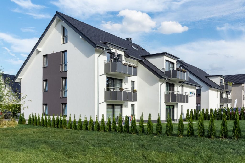 Modernes weißes Mehrfamilienhaus mit Balkonen und dunklem Dach, umgeben von gepflegten kleinen Nadelbäumen und grünem Gras, unter einem teilweise bewölkten blauen Himmel - fachmännisch ausgeführt vom Malerfachbetrieb Stein Maler.