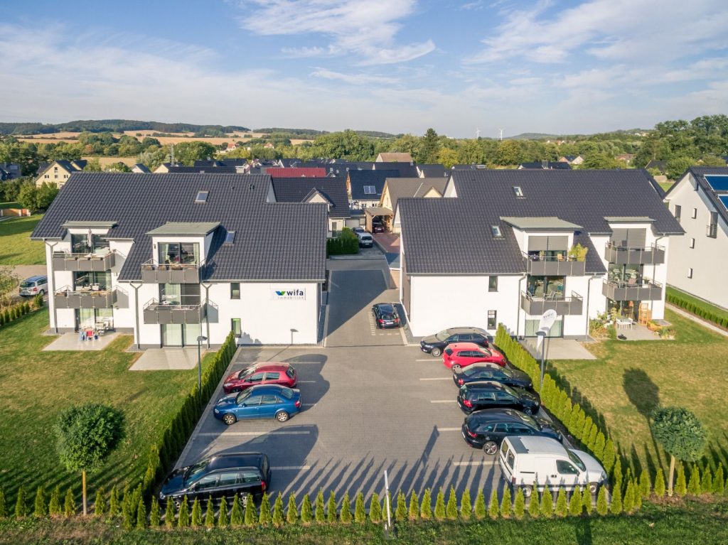 Luftaufnahme von modernen Wohnhäusern mit dunklen Dächern, die vom Malerfachbetrieb Stein fachmännisch bemalt wurden, Balkonen und einem mit Autos gefüllten Parkplatz; grüne Rasenflächen, Bäume und andere Häuser unter einem blauen Himmel mit vereinzelten Wolken.