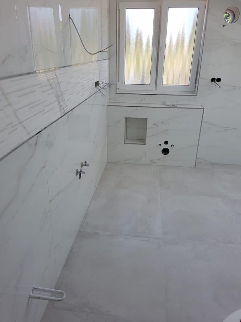 Unfertiges Bad mit weißen Marmorfliesen, freiliegenden Rohrleitungen, Elektrokabeln und einem kleinen Fenster; Waschbecken und Toilette sind noch nicht installiert - perfekt für den Malerfachbetrieb Stein aus Herford, der hier sein Können unter Beweis stellt.