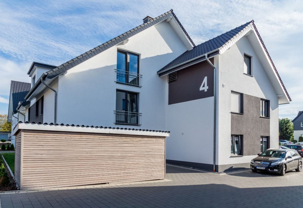 Modernes weißes zweistöckiges Gebäude mit dunkelgrauen Akzenten und der Nummer 4 an der Seite, neben einem hellen Holzschuppen. Fachmännisch gewartet vom Malerfachbetrieb Stein Maler. Vor dem Haus steht ein schwarzes Auto auf einer gepflasterten Einfahrt bei teilweise bewölktem Himmel.