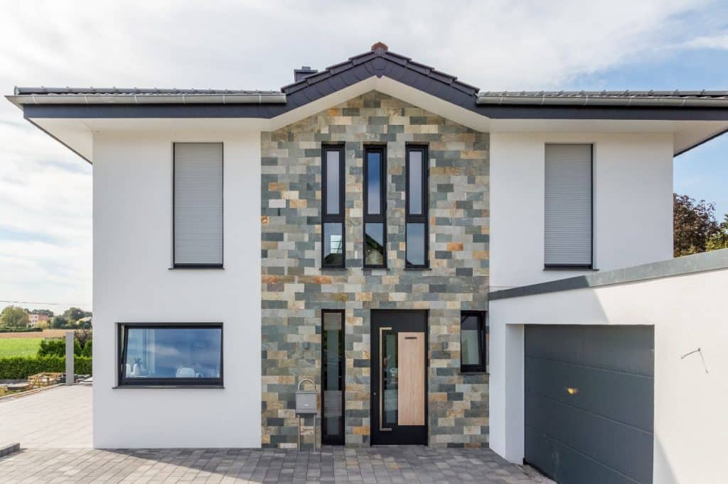 Modernes zweistöckiges Haus mit einer grauen Steinfassade von Malerfachbetrieb Stein, großen vertikalen Fenstern, weißen Wänden und einem grauen Garagentor. Die Einfahrt und der Eingang sind mit hellgrauen Ziegeln gepflastert, was für ein schlichtes, elegantes Aussehen sorgt.