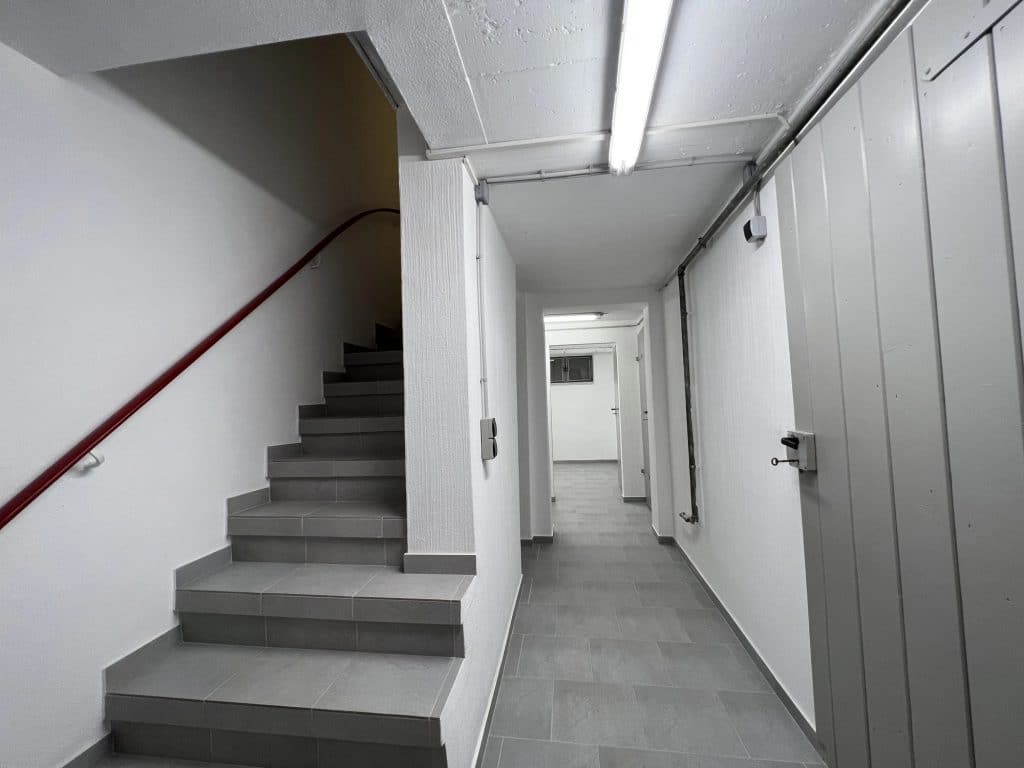 Ein sauberer, grau gefliester Flur in Herford mit geschlossenen Schranktüren auf der rechten Seite und einer Treppe mit rotem Handlauf auf der linken Seite - fachmännisch ausgeführt vom Malerfachbetrieb Stein, Ihrem Maler des Vertrauens. Leuchtstoffröhren erhellen die weißen Wände und den Boden.