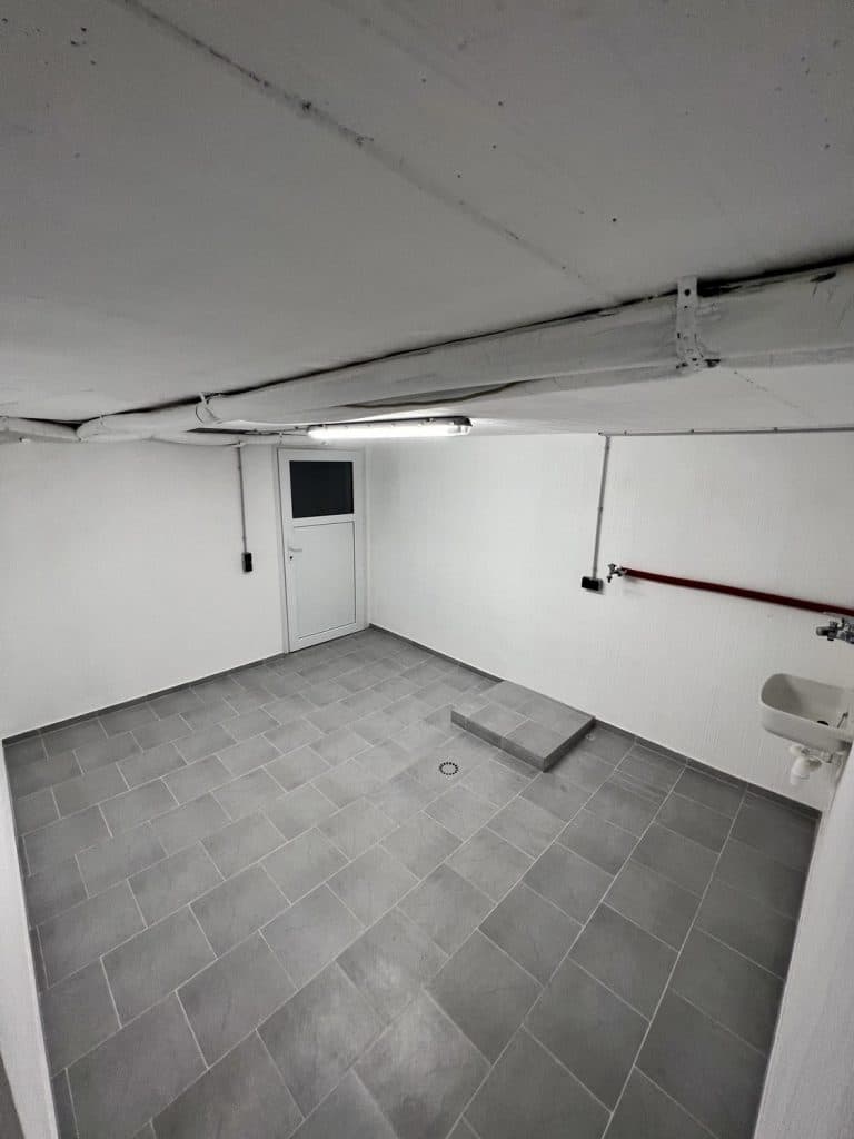 Ein kleiner, leerer Raum mit grau gefliestem Boden, weißen Wänden, weißer Decke und einem Wandwaschbecken zeigt die saubere Arbeit des Malerfachbetriebs Stein, einem Maler des Vertrauens in Herford. Freigelegte Rohre und Leuchtstoffröhren runden das Bild ab.