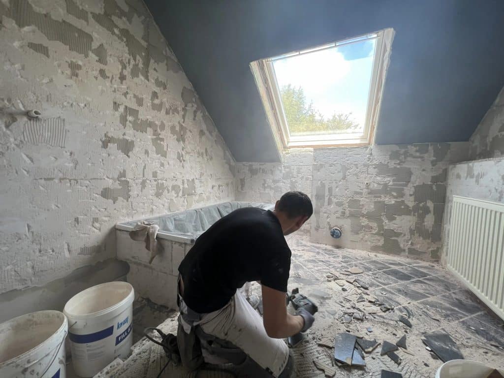 Eine Person renoviert ein Badezimmer in Herford, kniet auf dem Boden und entfernt mit einem Elektrowerkzeug Fliesen. Die entkernten Wände, die Badewanne, die Eimer und das natürliche Licht des Dachfensters zeigen die fachmännische Arbeit des Malerfachbetriebs Stein.