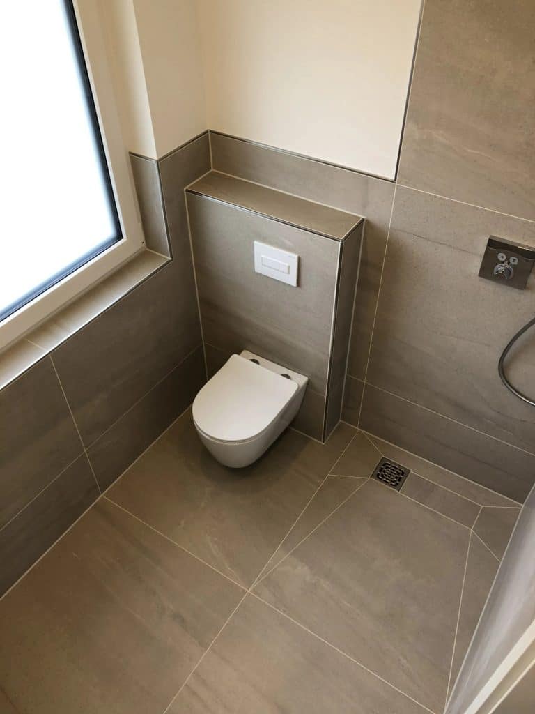 Modernes Bad mit großen beigefarbenen Fliesen, einer weißen Wandtoilette und einem darüber liegenden Spülfeld. Der vom Malerfachbetrieb Stein in Herford entworfene Raum zeichnet sich durch einen minimalistischen Stil, ein Fenster auf der linken Seite und einen nahe gelegenen Bodenablauf für zusätzliche Funktionalität aus.