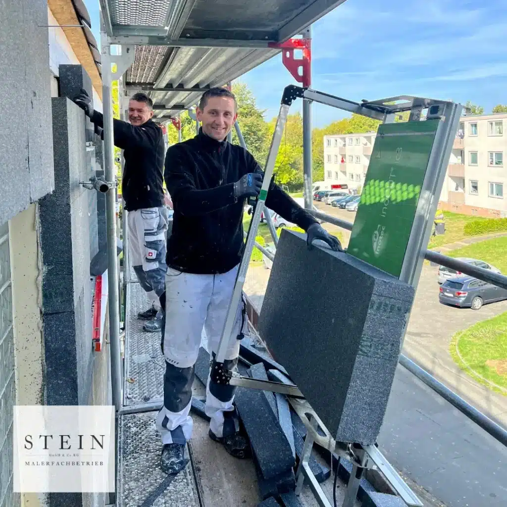 Zwei Bauarbeiter des Malerfachbetriebs Stein stehen auf einem Gerüst in Herford und lächeln, während sie Dämmplatten an der Außenfassade eines Gebäudes anbringen. Einer hält eine große Platte. Im Hintergrund sind Wohnhäuser und Grünanlagen zu sehen.