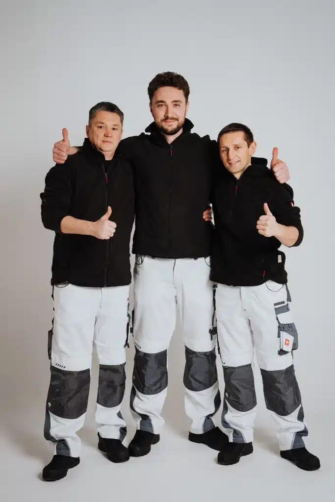 Drei Männer in passenden schwarzen Jacken und weißen Arbeitshosen stehen zusammen in einem Atelier, lächeln und zeigen den Daumen nach oben. Sie repräsentieren stolz den Malerfachbetrieb Stein, Ihr vertrauter Maler aus Herford. Der Mann in der Mitte hat einen Arm um jeden der anderen gelegt.