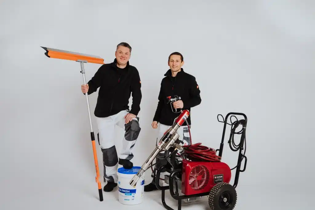 Zwei Männer in Arbeitsuniformen vom Malerfachbetrieb Stein in Herford posieren mit professionellen Reinigungsgeräten, darunter ein langstieliger Abzieher, ein Eimer, ein roter Hochdruckreiniger und Schläuche, vor einem schlichten weißen Hintergrund.