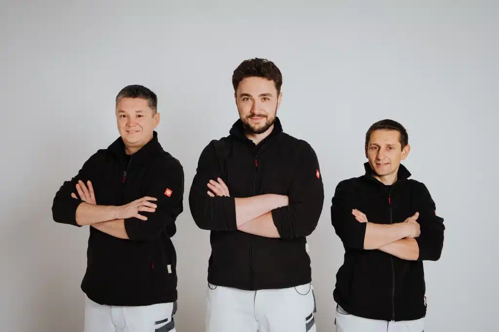 Drei Männer stehen nebeneinander vor einem schlichten weißen Hintergrund. Sie repräsentieren den Malerfachbetrieb Stein aus Herford, tragen alle schwarze Jacken und weiße Hosen, haben die Arme verschränkt und lächeln in die Kamera.