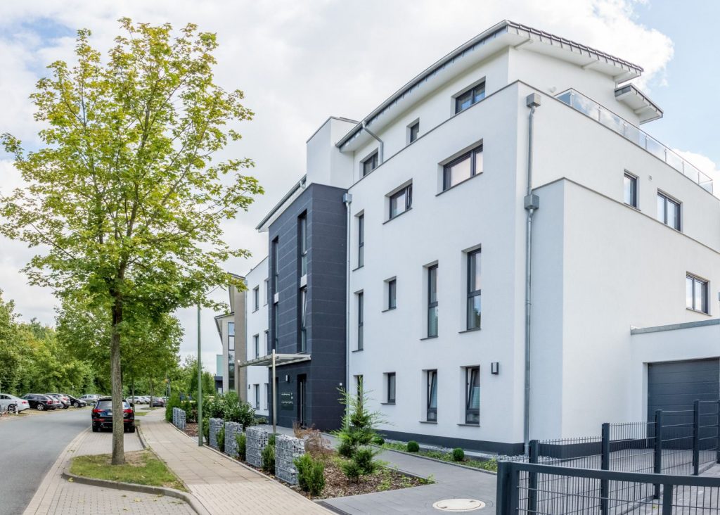 Ein modernes, weißes, dreistöckiges Wohnhaus mit großen Fenstern und Balkonen, das vom Malerfachbetrieb Stein fachmännisch fertiggestellt wurde, liegt an einer ruhigen, von Bäumen gesäumten Straße mit parkenden Autos und Grünflächen entlang des Gehwegs.