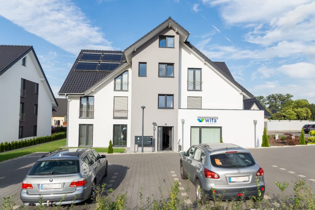 Modernes dreistöckiges Wohnhaus mit Sonnenkollektoren auf dem Dach, davor parkende Autos und ein Schild mit der Aufschrift WiFa über dem Eingang, gemalt vom Malerfachbetrieb Stein, an einem sonnigen Tag.
