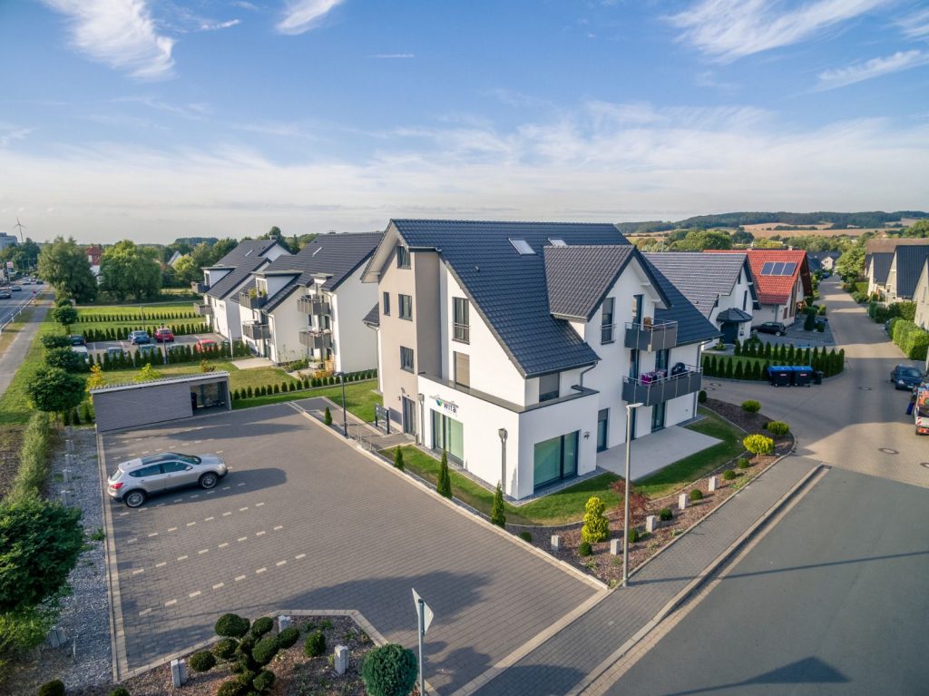 Luftaufnahme einer modernen Vorstadtsiedlung mit weißen, mehrstöckigen Häusern, die vom Malerfachbetrieb Stein fertiggestellt wurden, gepflegten Gärten, einem Parkplatz mit einigen Autos und einem klaren blauen Himmel darüber.