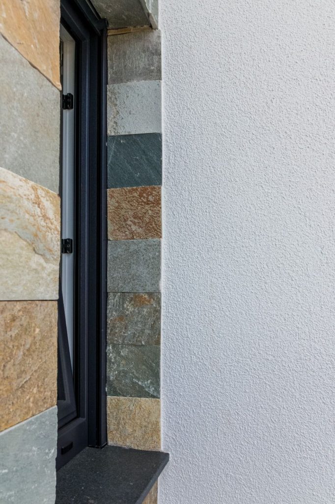 Nahaufnahme eines in eine Wand eingelassenen Fensters, bei dem der Malerfachbetrieb Stein mehrfarbige Steinfliesen auf der linken Seite mit einfarbigem weißem Strukturputz auf der rechten Seite verbindet und so die fachmännische Handwerkskunst von Maler zeigt.