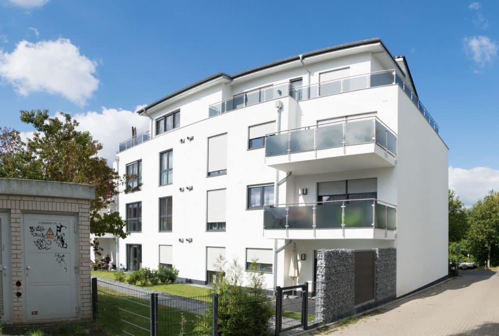 Modernes weißes Mehrfamilienhaus mit großen Fenstern und Balkonen, fachmännisch vom Malerfachbetrieb Stein bemalt, an einer Ecke mit einer kleinen grünen Wiese, Bäumen und einem gepflasterten Bürgersteig unter einem strahlend blauen Himmel mit vereinzelten Wolken.