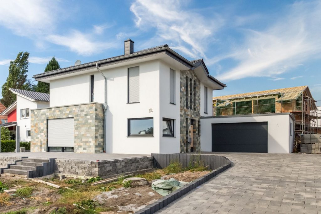 Modernes zweistöckiges Haus mit weißen Wänden, gemusterten Steinakzenten von Malerfachbetrieb Stein, großen Fenstern mit geschlossenen Rollläden und einer Doppelgarage. Die gepflasterte Auffahrt führt zu einer noch nicht fertig gestellten Gartenanlage; ein Nachbarhaus befindet sich im Bau.