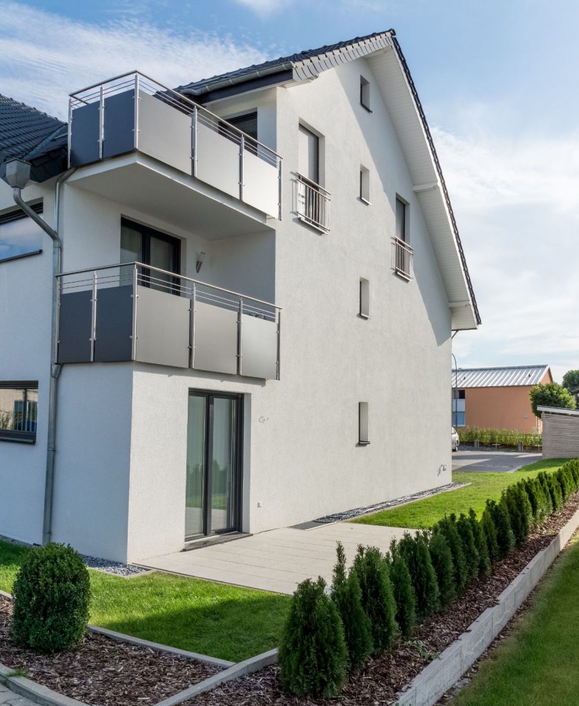 Modernes, weißes, zweistöckiges Haus vom Malerfachbetrieb Stein, mit zwei grauen Metallbalkonen, großen Fenstern, einer kleinen Terrasse, ordentlich getrimmtem Rasen und einer Reihe von Sträuchern entlang des Gehwegs unter blauem Himmel.