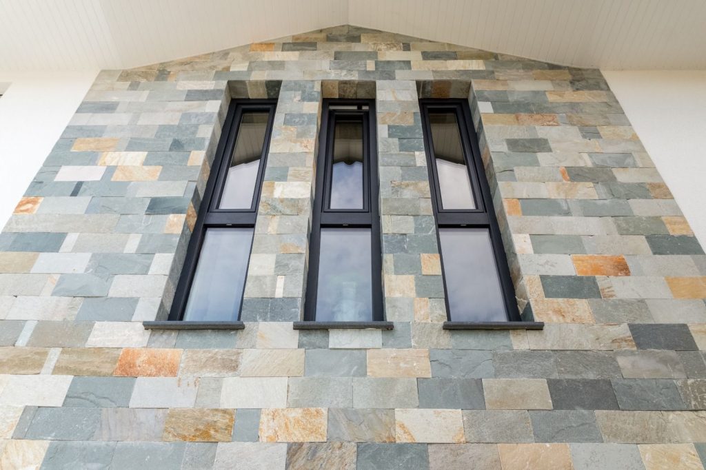 Eine moderne, vom Malerfachbetrieb Stein entworfene Fassade mit mehrfarbigen, rechteckigen Steinplatten und drei hohen, schmalen, vertikal angeordneten, schwarz gerahmten Fenstern.