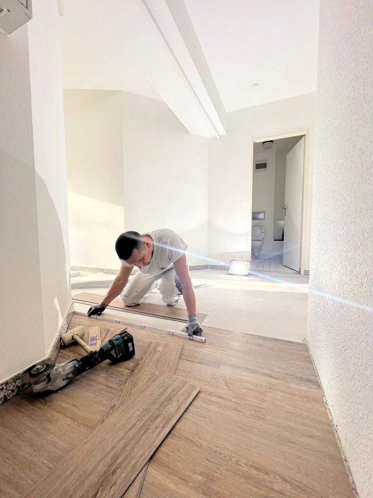 Eine Person kniet auf dem Boden und verlegt Vinyl-Dielen in Holzoptik in einem hellen Raum. Werkzeuge und Bodenbelagsmaterialien des Malerfachbetriebs Stein liegen in der Nähe verstreut, während natürliches Licht den Raum erfüllt. Im Hintergrund sind eine offene Tür und eine Toilette zu sehen.