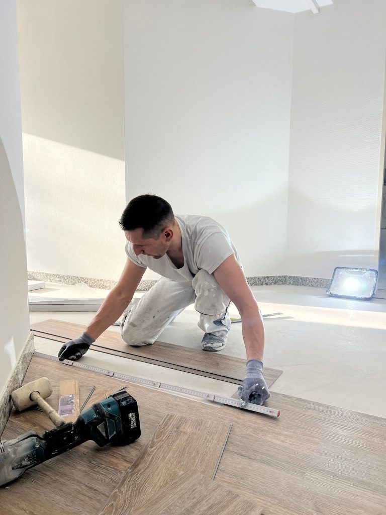 Ein Mann vom Malerfachbetrieb Stein, bekleidet mit Arbeitskleidung und Handschuhen, misst kniend ein Stück Bodenbelag mit einem Lineal aus; Werkzeuge und eine Lampe stehen in der Nähe, während das Sonnenlicht den Raum erfüllt.