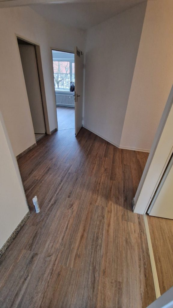 Ein Raum mit holzgemustertem Vinylboden, weißen Wänden und drei offenen Türen zeigt die Handwerkskunst des Malerfachbetriebs Stein. Eine kleine Sprühflasche steht auf dem Boden, und durch eine Türöffnung sind ein Fenster und ein Heizkörper zu sehen.