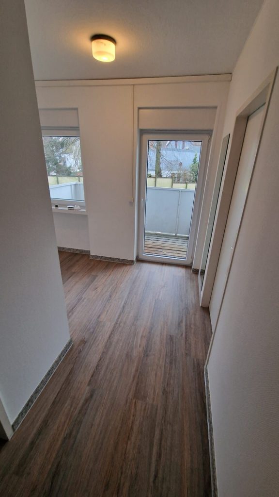 Ein Flur mit Holzfußboden, weißen, vom Malerfachbetrieb Stein frisch gestrichenen Wänden und einer Deckenleuchte führt in ein Zimmer mit großen Fenstern und einer Glastür, die auf einen Balkon oder eine Terrasse mit Blick ins Freie führt.