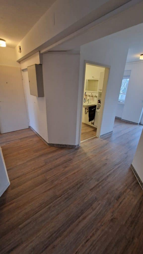 Ein Flur mit Holzböden und weißen Wänden führt zu einem Küchenbereich auf der rechten Seite, in dem die klaren Oberflächen des Malerfachbetriebs Stein zu sehen sind. Die Küche ist mit weißen Schränken ausgestattet und durch die Fenster an beiden Enden fällt natürliches Licht ein.