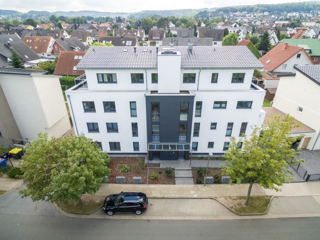 Luftaufnahme eines modernen weißen Mehrfamilienhauses mit grauem Dach, professionell gemalt von Malerfachbetrieb Stein, umgeben von Bäumen und geparkten Autos in einem Wohnviertel mit grünen Hügeln im Hintergrund.