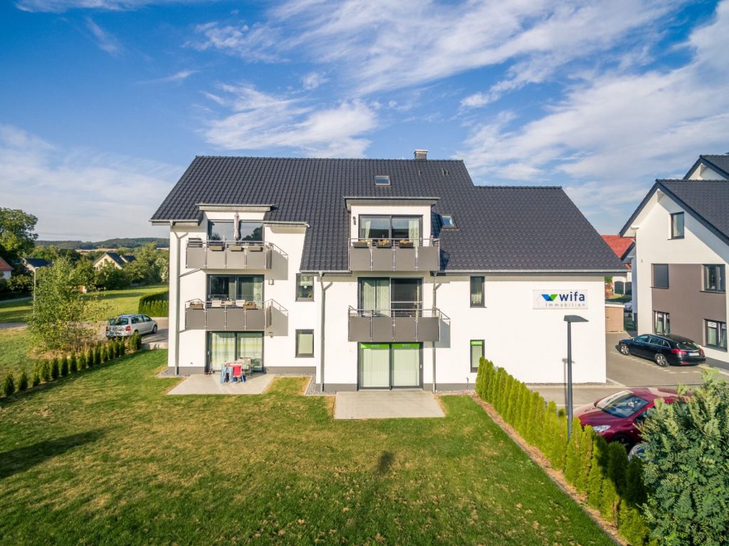 Ein modernes dreistöckiges Mehrfamilienhaus mit Balkonen, weißen, vom Malerfachbetrieb Stein frisch verputzten Außenwänden und einem dunklen Satteldach steht auf einer gepflegten Rasenfläche. Ein wifa-Schild ist vorhanden, im Hintergrund sind die Nachbarhäuser zu sehen.