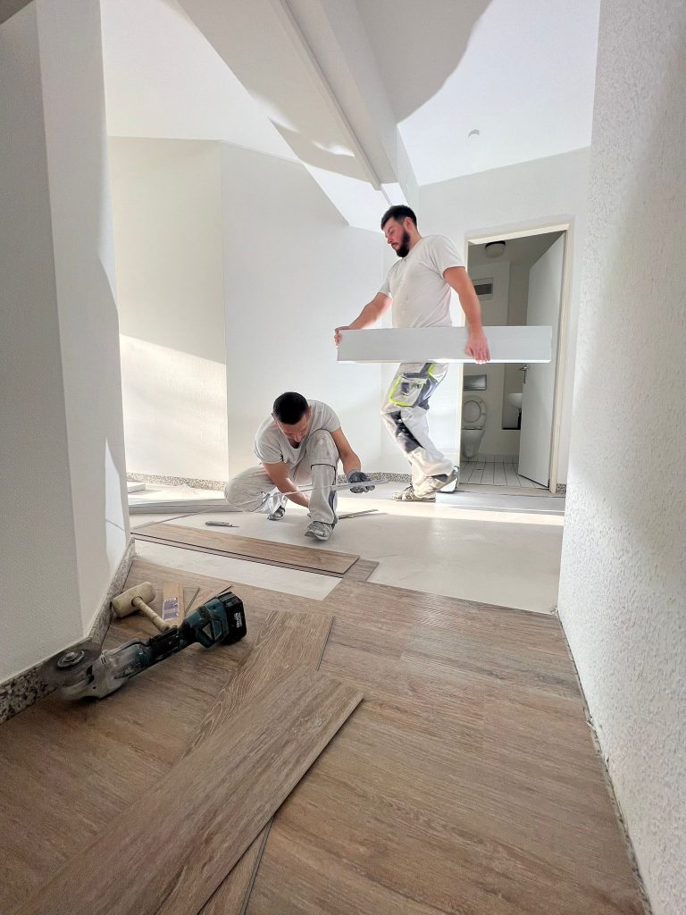 Zwei Arbeiter des Malerfachbetriebs Stein verlegen einen Bodenbelag in einem hellen Raum. Einer kniet und richtet Holzbretter aus, während der andere ein langes Brett hält. Werkzeuge und Bodenbelagsmaterialien liegen verstreut auf dem Boden.