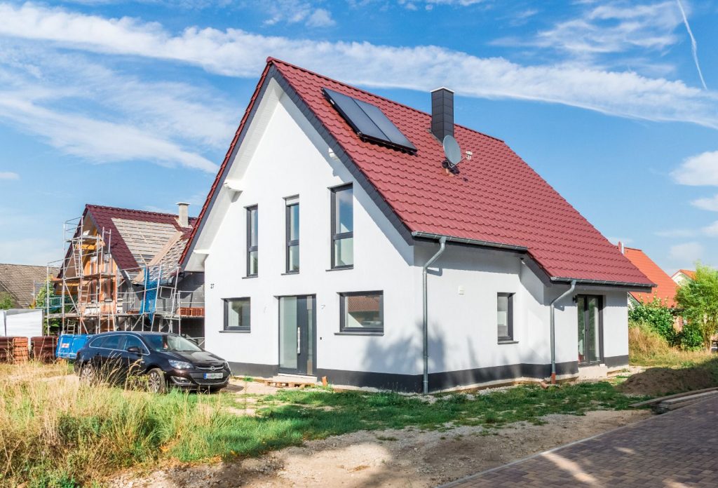 Ein modernes weißes Haus mit rotem Satteldach, Sonnenkollektoren und großen Fenstern steht an einem sonnigen Tag da. Es wurde vom Malerfachbetrieb Stein fertiggestellt und ist ein Musterbeispiel für fachmännische Malerarbeit. Draußen parkt ein schwarzes Auto, in der Nähe wird gerade ein weiteres Haus gebaut.