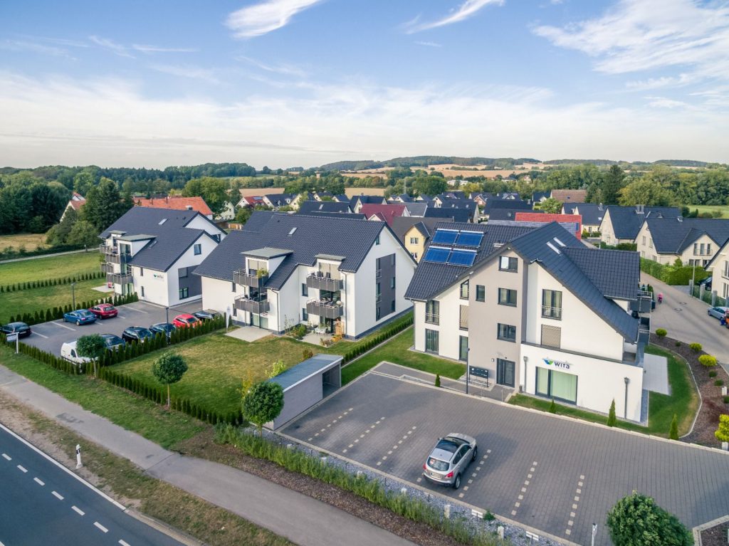 Luftaufnahme eines modernen Wohnviertels mit weißen und grauen mehrstöckigen Häusern des Malerfachbetriebs Stein, ordentlich geparkten Autos, grünen Rasenflächen, Bäumen und umliegender Landschaft unter blauem Himmel.
