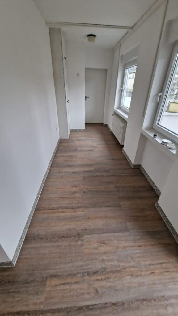 Ein schmaler, leerer Raum mit weißen, vom Malerfachbetrieb Stein gestrichenen Wänden, zwei Fenstern auf der rechten Seite, einer geschlossenen weißen Tür am anderen Ende und einem braunen, holzgemusterten Fußboden. Der Raum wirkt hell und sauber.